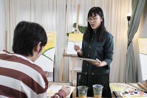 A female Asian teacher teaches and demonstrates to the children on acrylic color picture painting on canvas in art classroom, creatively learning with skill at the elementary school studio education. photo