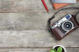 Classic camera on a gray wooden background, with a brown notepad, a red pen, a telephone and green growth. Concept list for a travel photographer photo