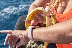 Latin woman, middle-aged, resting, regaining strength, eating, drinking water, after a gym session, burning calories, keeping fit, outdoors by the sea, wearing headphones and smart watch photo