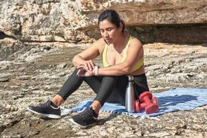 Latin woman, middle-aged, resting, regaining strength, eating, drinking water, after a gym session, burning calories, keeping fit, outdoors by the sea, wearing headphones and smart watch photo