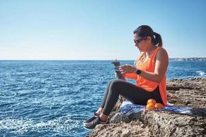 Latin woman, middle-aged, resting, regaining strength, eating, drinking water, after a gym session, burning calories, keeping fit, outdoors by the sea, wearing headphones and smart watch photo