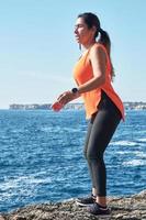 FITNESS LATIN WOMAN IN SPORTS SET TRAINING WITH ELASTIC BAND, WEIGHTS, GYM EXERCISES, IN FRONT OF THE WATER. photo