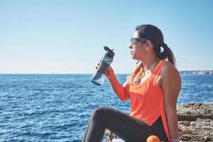 Latin woman, middle-aged, resting, regaining strength, eating, drinking water, after a gym session, burning calories, keeping fit, outdoors by the sea, wearing headphones and smart watch photo