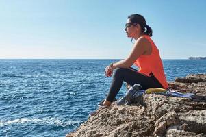 Latin woman, middle-aged, resting, regaining strength, eating, drinking water, after a gym session, burning calories, keeping fit, outdoors by the sea, wearing headphones and smart watch photo