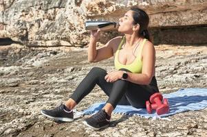 seated woman, wearing sports clothes, drinking water from a thermal bottle, next to a pair of dumbbells, photo