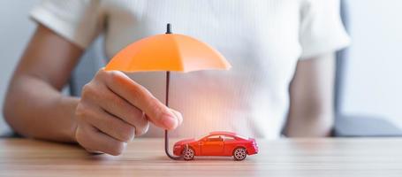 mano de mujer de negocios sosteniendo un paraguas naranja y cubriendo el juguete del coche rojo en la mesa. seguro de automóvil, garantía, reparación, concepto financiero, bancario y monetario foto