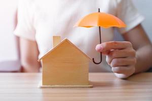Businesswoman hand holding orange Umbrella cover wooden Home model. real estate, insurance and property concepts photo