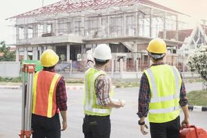 back view group of engineer, technician and architect with safety helmet planning about building plan with blueprint in modern city building background, construction site, industry and worker concept photo