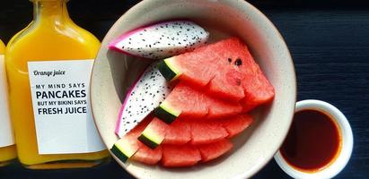 vista superior de rodajas frescas de sandía y fruta de dragón en un plato con una botella de jugo de naranja en una mesa de madera negra. endecha plana de fruta fresca en un plato sobre fondo de madera. conjunto de alimentos saludables y dieta foto