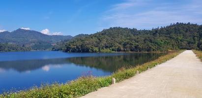 Road or street beside lake or river with green mountain and blue sky background at Huai Bon reservoir Chiang Mai, Thailand. Among nature with reflection on water and copy space. Beauty in nature