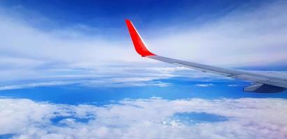 Red and gray or grey wing of plane with blue sky and white cloud background and copy space. Aerial view of airplane flying and going to destination. Top view of vehicle travel on high place. Transport photo