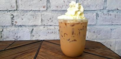 Plastic glass of iced coffee with whipped cream on top on brown wooden table with white rough wall background. Cold espresso, latte or cappuccino at caf. Drinking and food concept with copy space. photo