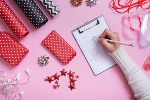 lista de nombres y envolver regalos de navidad planos sobre fondo rosa. vista superior del concepto de preparación navideña. foto