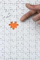 The texture of a white jigsaw puzzle in the assembled state with one missing element, forming an orange space, pointed to by the finger of the male hand photo