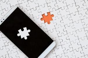 A modern big smartphone with several puzzle elements on the touch screen lies on a white jigsaw puzzle in an assembled state with missing elements photo