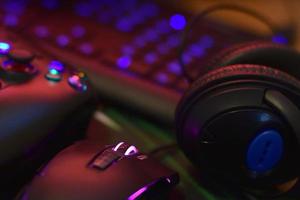 Modern gamepad and gaming mouse lies with keyboard and headphones on table in dark playroom scene. Electronic devices and technology concept photo