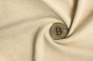 The golden bitcoin lies on a blanket made of soft and fluffy light orange fleece fabric with a large number of relief folds. The shape of the folds resembles a fan from a video card cooler photo