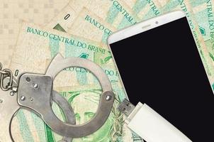 1 Brazilian real bills and smartphone with police handcuffs. Concept of hackers phishing attacks, illegal scam or malware soft distribution photo