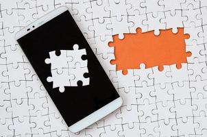 A modern big smartphone with several puzzle elements on the touch screen lies on a white jigsaw puzzle in an assembled state with missing elements photo