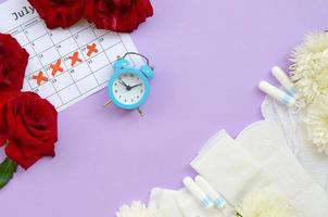 Menstrual pads and tampons on menstruation period calendar with blue alarm clock and red rose flowers photo