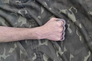 Male fist with brass knuckles on the background of a camouflage jacket. The concept of skinhead culture, handmade melee weapons photo