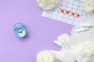 Menstrual pads and tampons on menstruation period calendar with blue alarm clock and white flowers photo