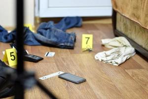 investigación de la escena del crimen - numeración de evidencias después del asesinato en el apartamento. teléfono inteligente roto, billetera y ropa con marcadores de evidencia foto