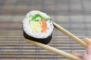 una mano con palillos sostiene un rollo de sushi sobre un fondo de estera de paja de bambú. comida asiática tradicional foto