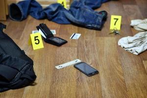 investigación de la escena del crimen - numeración de evidencias después del asesinato en el apartamento. teléfono inteligente roto, billetera y ropa con marcadores de evidencia foto