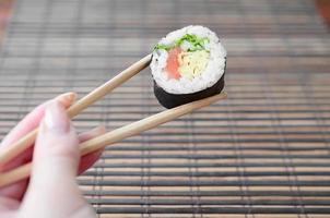 A hand with chopsticks holds a sushi roll on a bamboo straw serwing mat background. Traditional Asian food photo