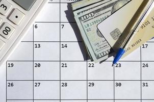 Pen and credit card on many hundred US dollar bills and calculator on calendar page close up photo