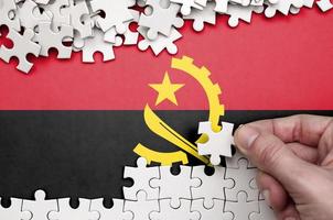 Angola flag  is depicted on a table on which the human hand folds a puzzle of white color photo