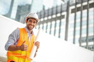 Young caucasioan man holding a big paper, guy wearing light blue shirt and jeans with orange vest and white helmet for security in construction area. photo