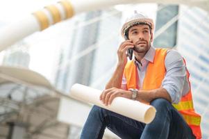 el ingeniero caucásico usa un teléfono inteligente para hablar, usa un chaleco naranja y un gran sombrero duro, y la otra mano sostiene el plano blanco en el sitio de trabajo del centro de la ciudad. foto