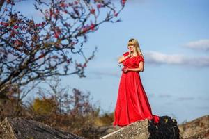 chica vestida de rojo sobre roca o estructura en ruinas de hormigón con serbal sobre fondo de otoño foto