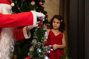 niña en vestido rojo decora un árbol de navidad con santa claus en casa foto
