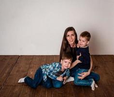 Happy mom with two children, in casual clothes, are photographed sitting on the floor. Photoshoot of a young mother with 2 sons, for white background. Warm family relationships. photo