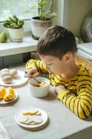 niño desayuna por la mañana con cereales con leche. desayuno de la mañana en la cocina antes de la escuela. el niño está comiendo en la mesa junto a la ventana. foto