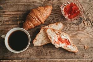 cut croissant with butter and strawberry jam with a cup of coffee, on a wooden background, top view. photo