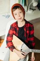 niño con un sombrero rojo y una camisa a cuadros toca la balalaika. chico guapo sosteniendo su guitarra. clases de música en casa. pasatiempo para el alma. música de enseñanza en casa foto