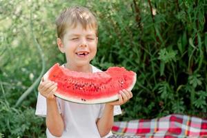 toothless little man eats watermelon white t-shirt. Picnic with watermelons. Summer mood. The boy is holding a large piece of watermelon in his hands. photo