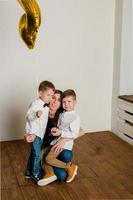 Happy mom with two children, in casual clothes, are photographed sitting on the floor. Photoshoot of a young mother with 2 sons, for white background. Warm family relationships. photo