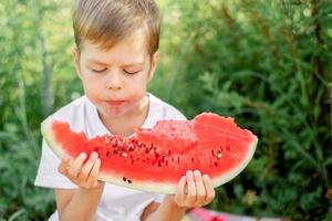 boy eating watermelon white t-shirt. Picnic with watermelons. The boy is holding a large piece of watermelon in his hands. Bright red juicy watermelon. photo