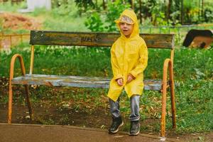 a boy in a yellow raincoat sits on a bench in the rain. A lonely child is lost on the street. Children without supervision. Bright clothes for children for a walk. photo