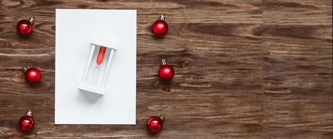 banner hourglass on a blank sheet of paper and and red christmas balls on a wooden background. Top view, flat lay photo