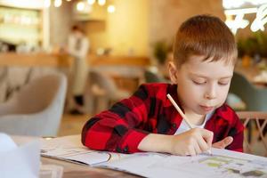 un niño guapo dibuja en la mesa con un lápiz. esperando un pedido en un café. el niño dibuja en páginas para colorear en un café. foto