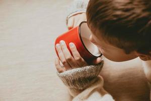 hands in a sweater hold hot cocoa, in a red mug, top view. A cozy photo with a mug in hand with copy space. Child drinks hot chocolate