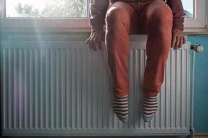 Man warms feet near a slightly warm radiator. Concept of energy crisis and increase in heating costs photo