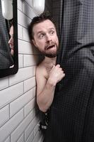 Wet, frightened man peeps out from behind the shower curtain and looks uneasy with a strange expression on his face. photo