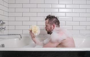 Sad man bathes in a bath with lush soap suds, he holds a sponge in his hands and looks thoughtfully. photo